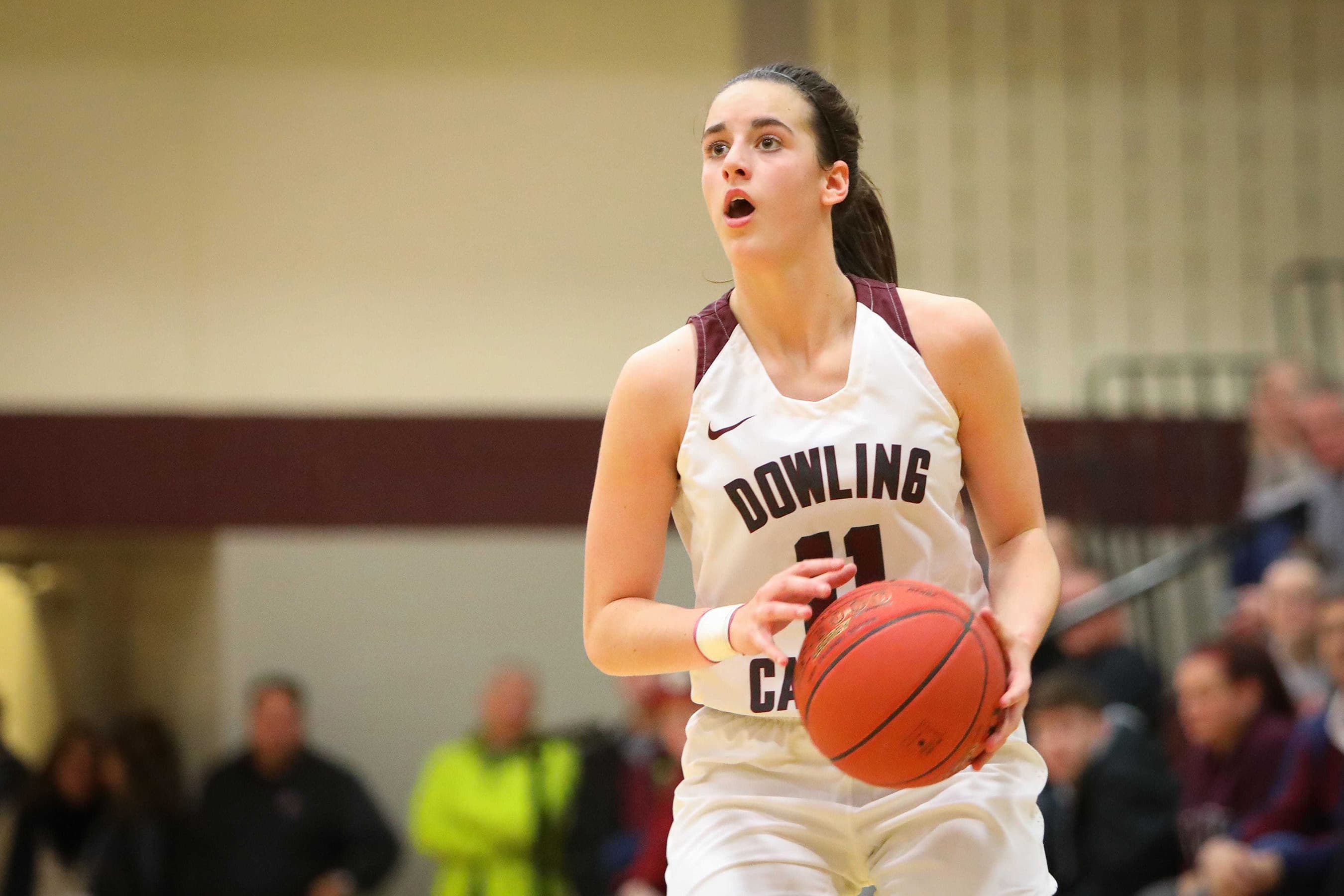 Basketball player in a white "Dowling" jersey holds a ball, looking focused. Indoor court setting with blurred spectators in the background.